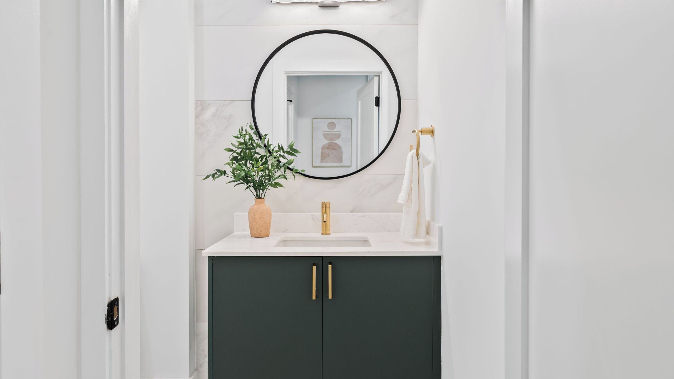 A bathroom with a white sink, dark cabinets, a gold faucet, a mirror with a black frame, and a potted plant.