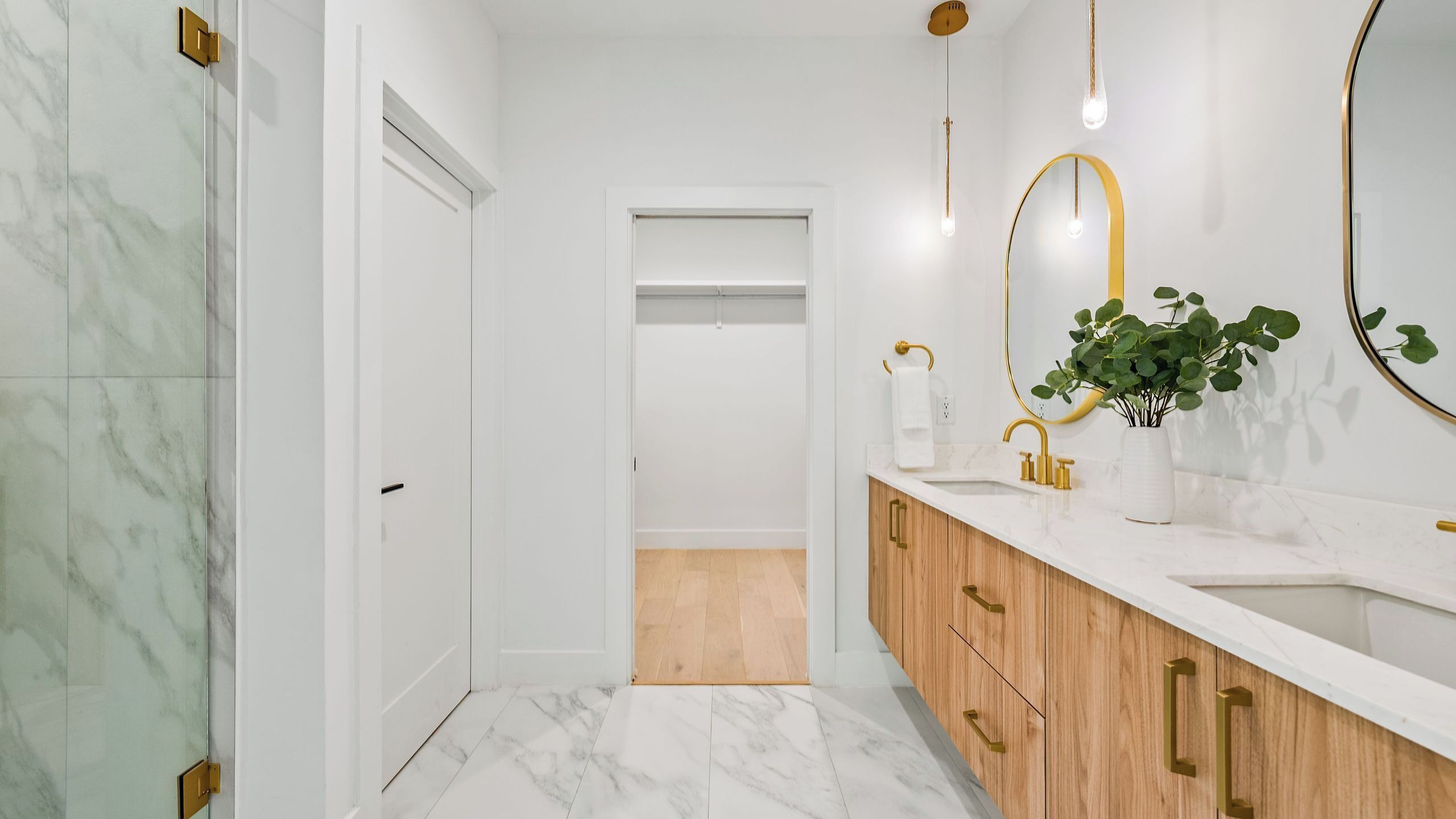 A modern bathroom with marble flooring, a wooden vanity, a white sink, and a large mirror with a gold frame.