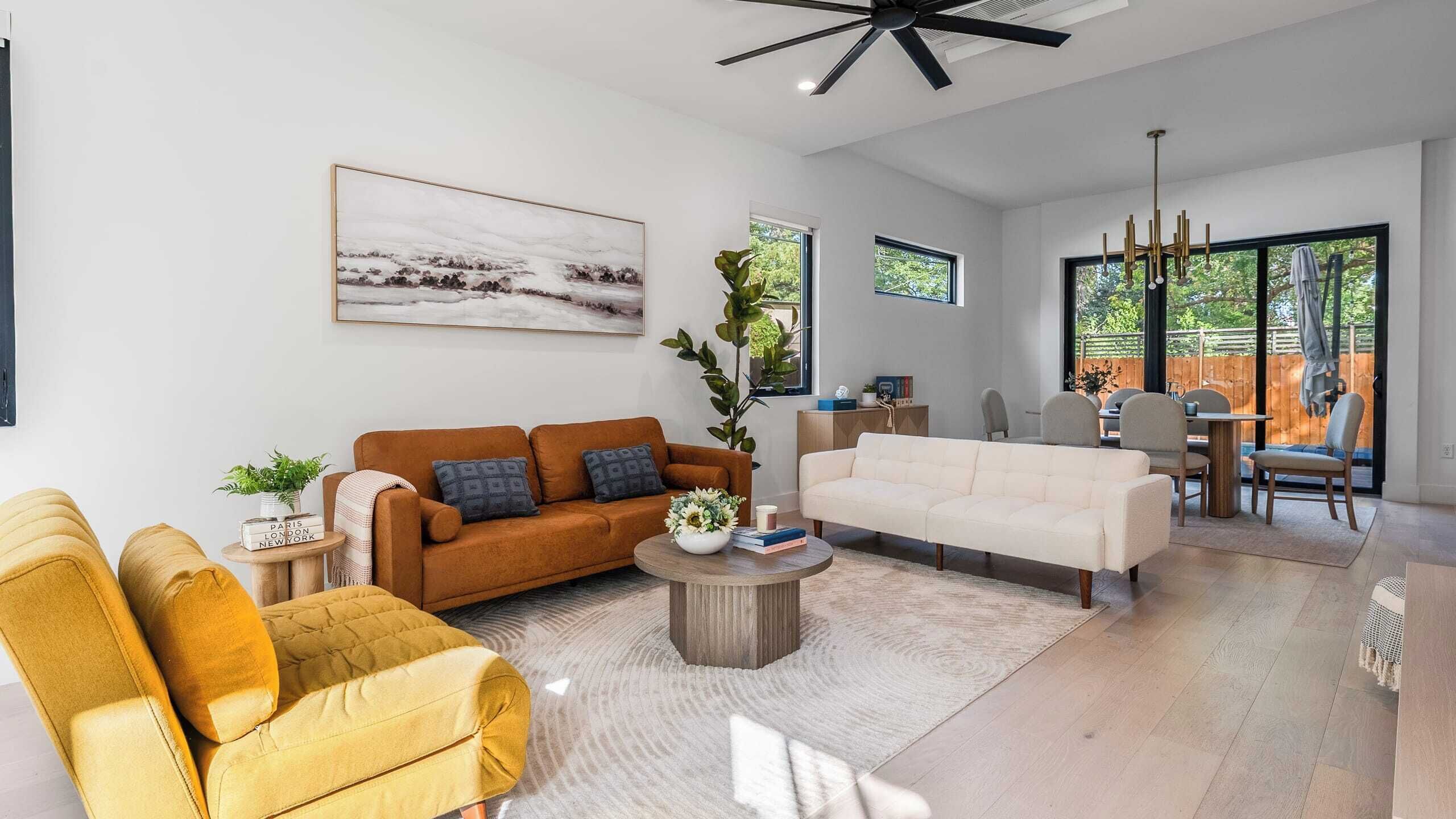 A spacious living room with a modern sofa, a yellow armchair, and a coffee table. There is a large window providing natural light and a view of the outdoors.