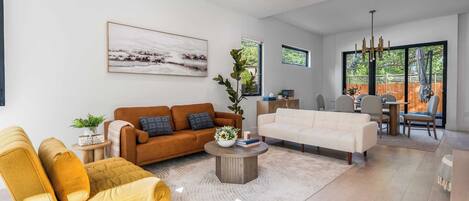 A spacious living room with a modern sofa, a yellow armchair, and a coffee table. There is a large window providing natural light and a view of the outdoors.