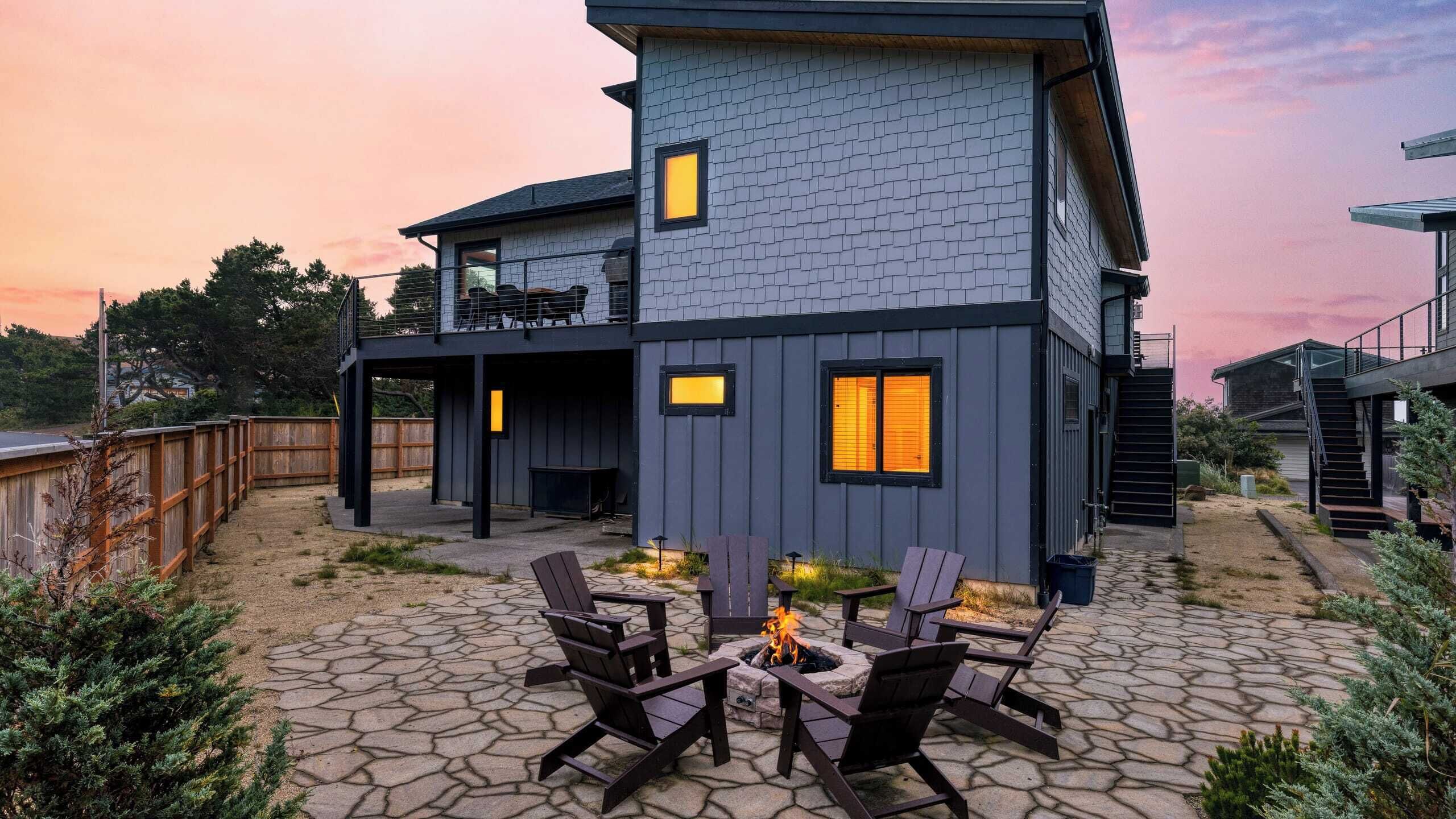 Una casa moderna de dos pisos con un patio de piedra, mobiliario exterior y un pozo de fuego.