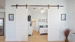 Ein Zimmer mit einer Schiebetür zu einem weiteren Raum, einem Bett mit Kissen, einem Fernseher auf einem Schrank und einer Vase mit Trockenpflanzen.