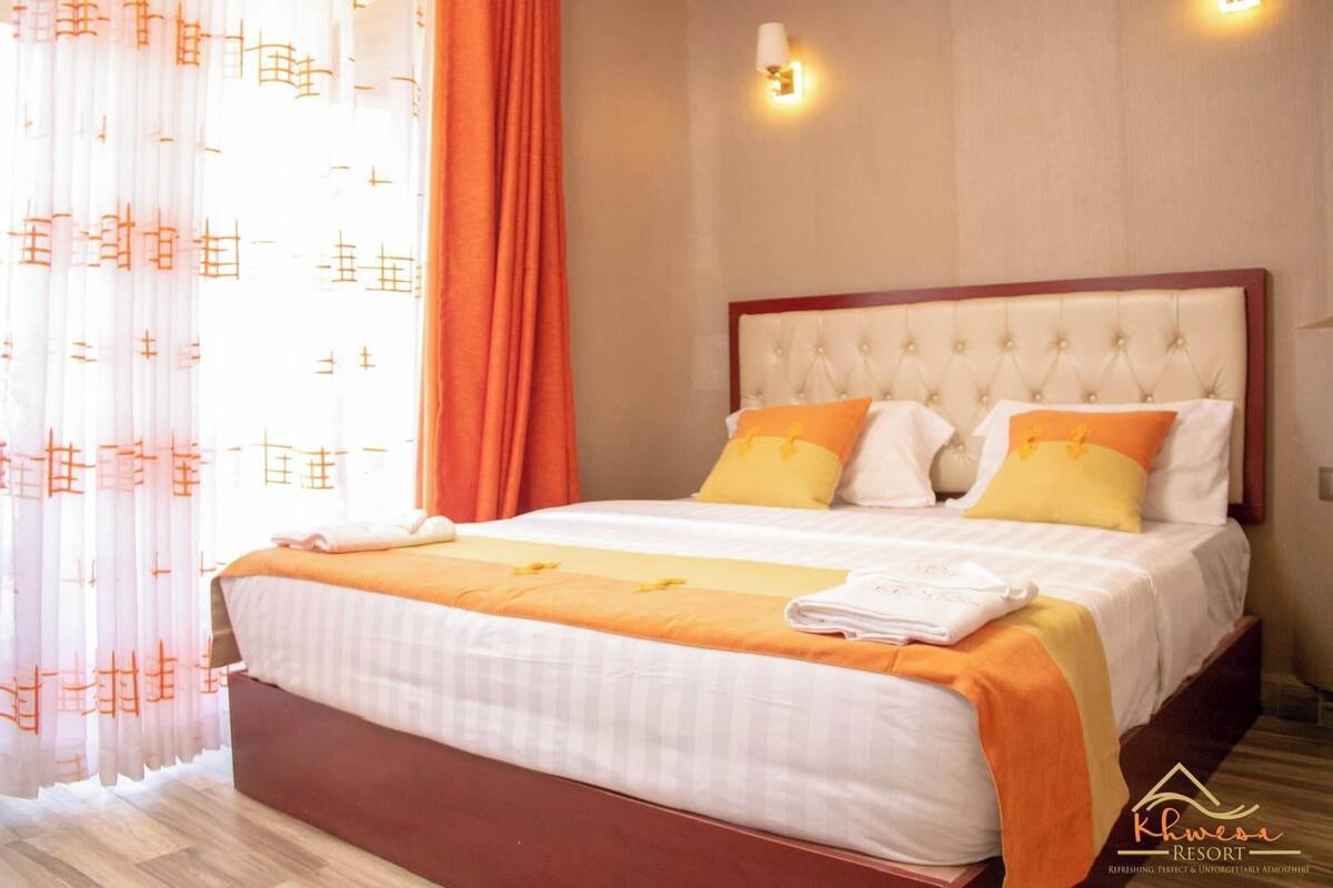 Una camera d'albergo con un letto grande, tende arancioni e una lampada fissata al muro.