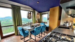 A dining area with a table set for four, a kitchen with a gas stove, and a view of green fields through large windows. - Casa Olanda (mornico losana)