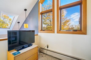Una habitación con una televisión sobre un mueble de madera, una lámpara colgante y dos ventanas que dan a árboles.
