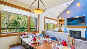 Un comedor con una mesa de madera dispuesta para cuatro personas, ventanas amplias con vista a árboles, un sofá y una chimenea.