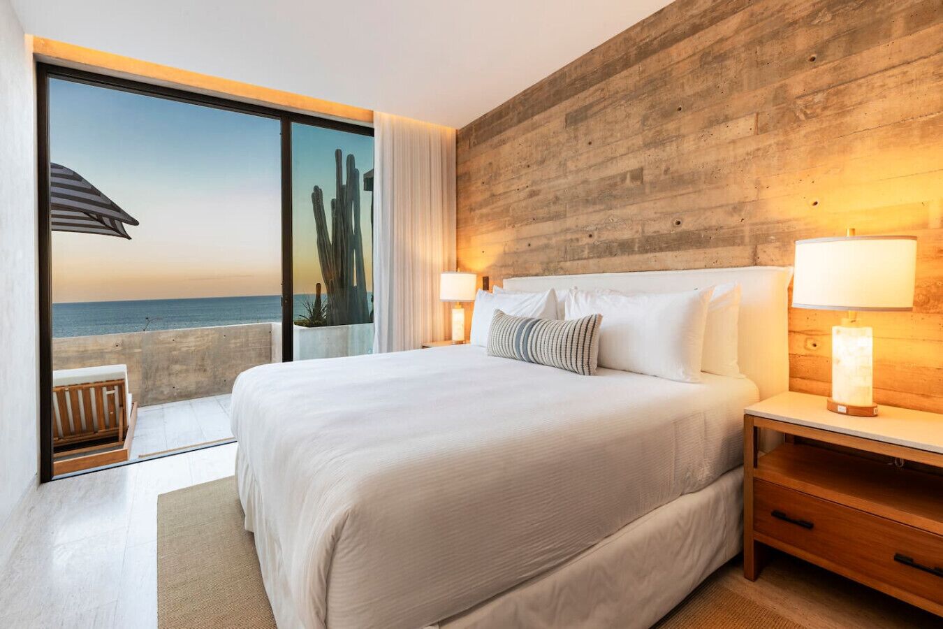 A bedroom with a large bed, a wooden headboard, a nightstand, and a view of the ocean through a sliding door.