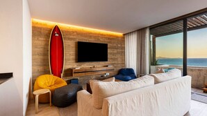 Una sala de estar moderna con un televisor de pantalla plana grande, una tabla de surf roja y vistas al mar a través de amplias ventanas.
