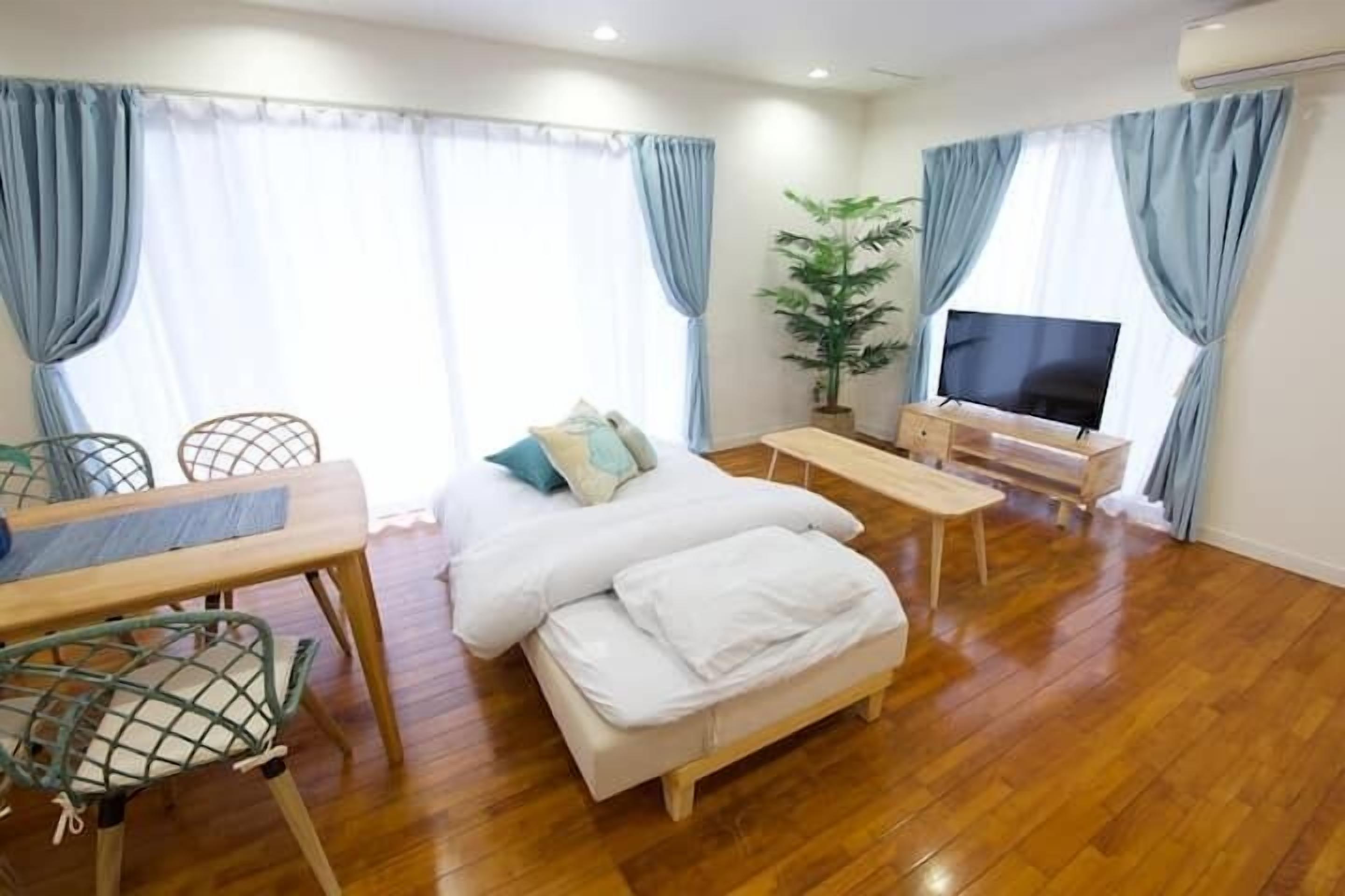 A cozy hotel room with a wooden floor, a bed with white bedding, a TV, a desk with a chair, and a potted plant.