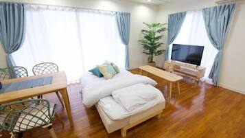 A cozy hotel room with a wooden floor, a bed with white bedding, a TV, a desk with a chair, and a potted plant.