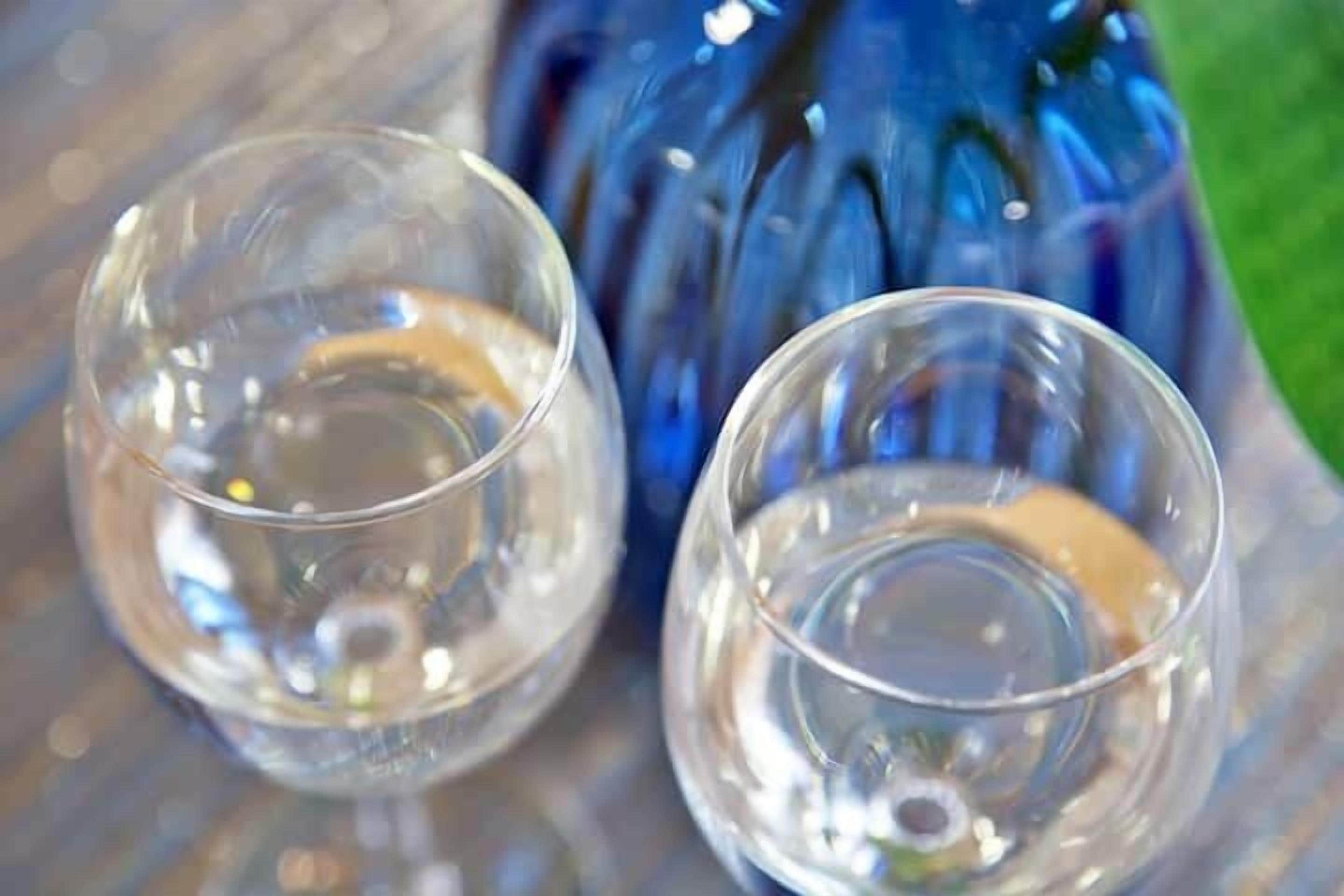Two clear glasses on a reflective surface with a blue bottle in the background.