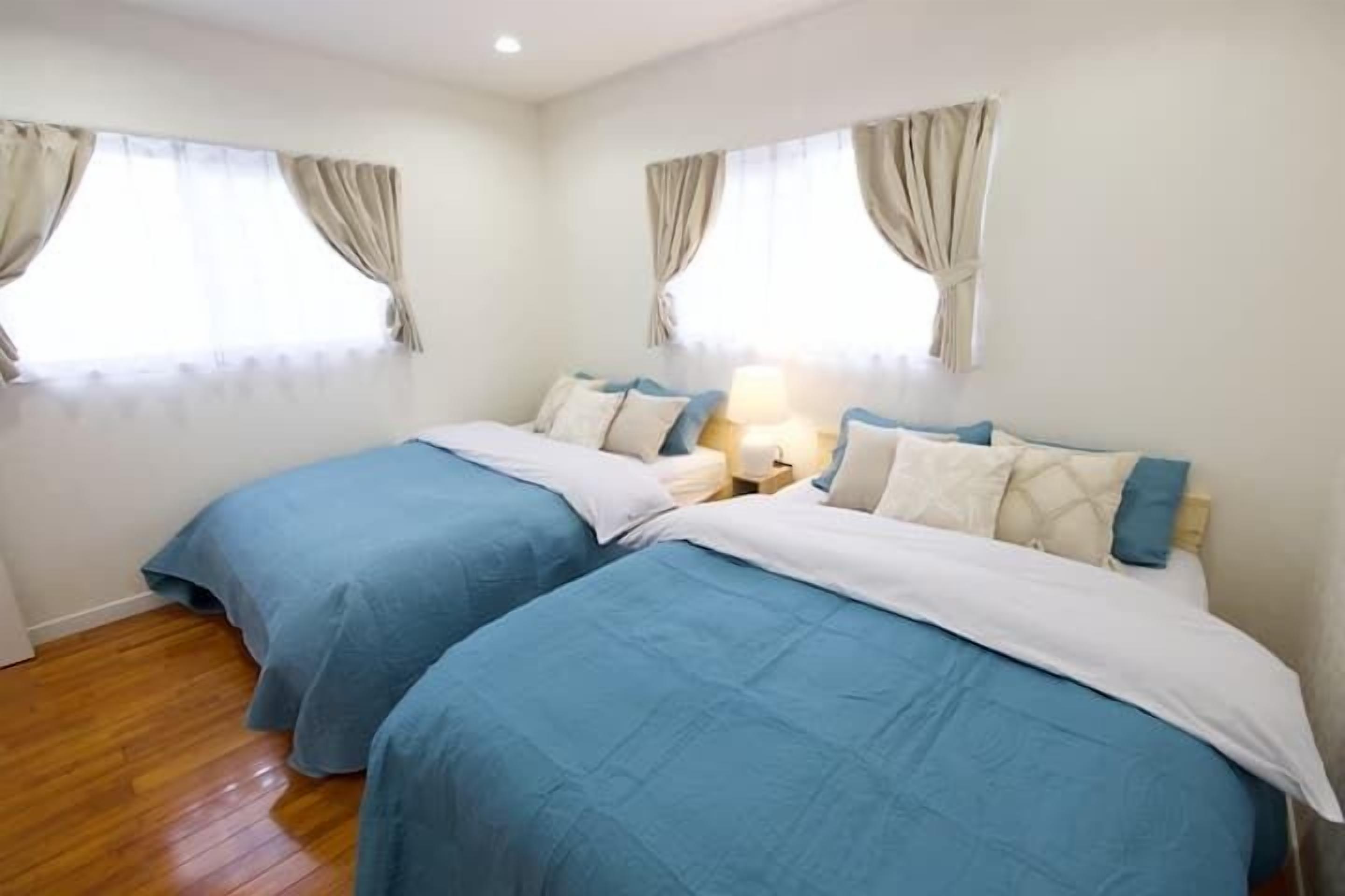 A hotel room with two beds, beige curtains, a wooden floor, and a nightstand with a lamp.