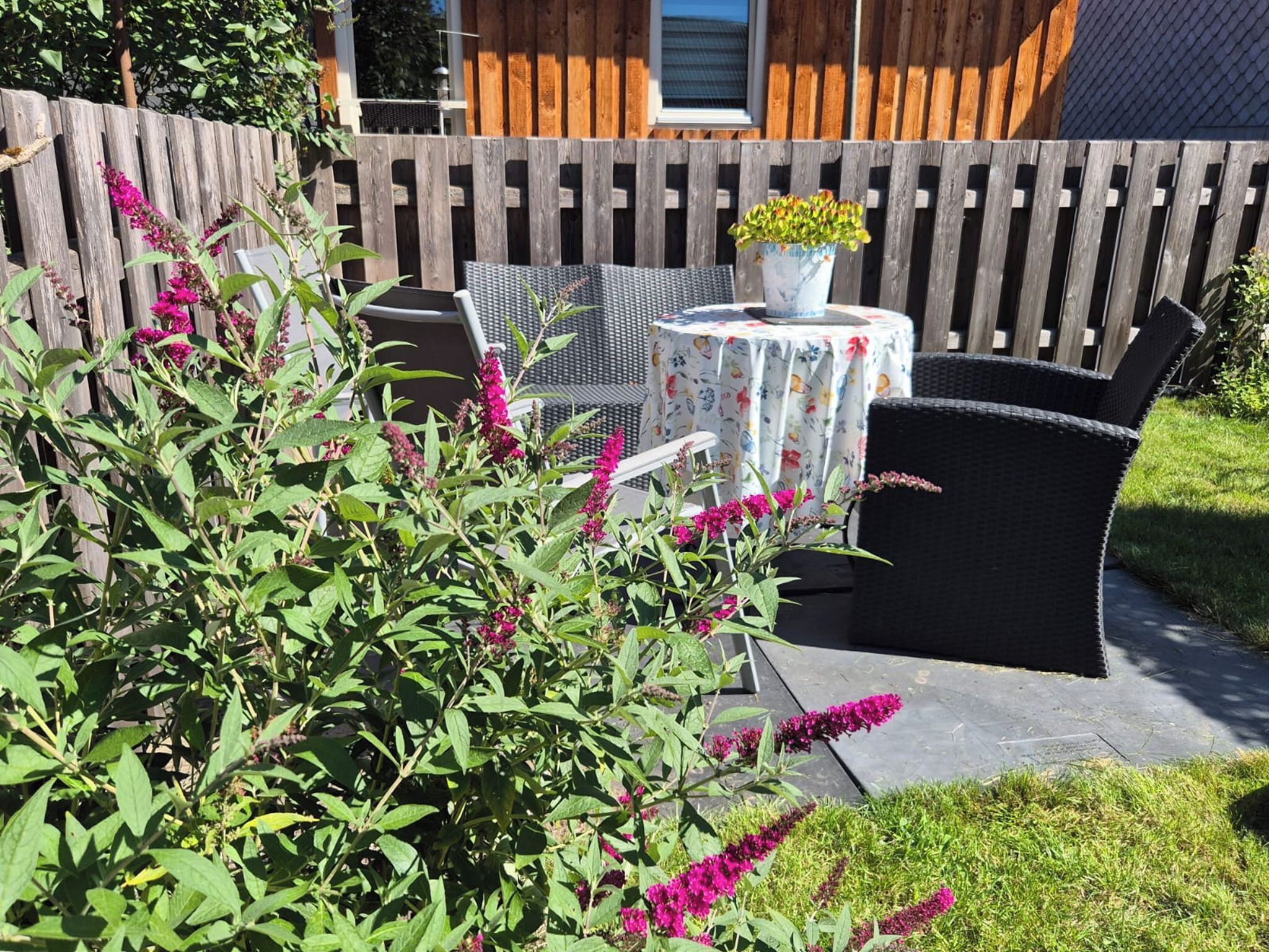 Een terras buiten met een houten omheining, een ronde tafel met een bloemen tafelkleed en rieten stoelen.