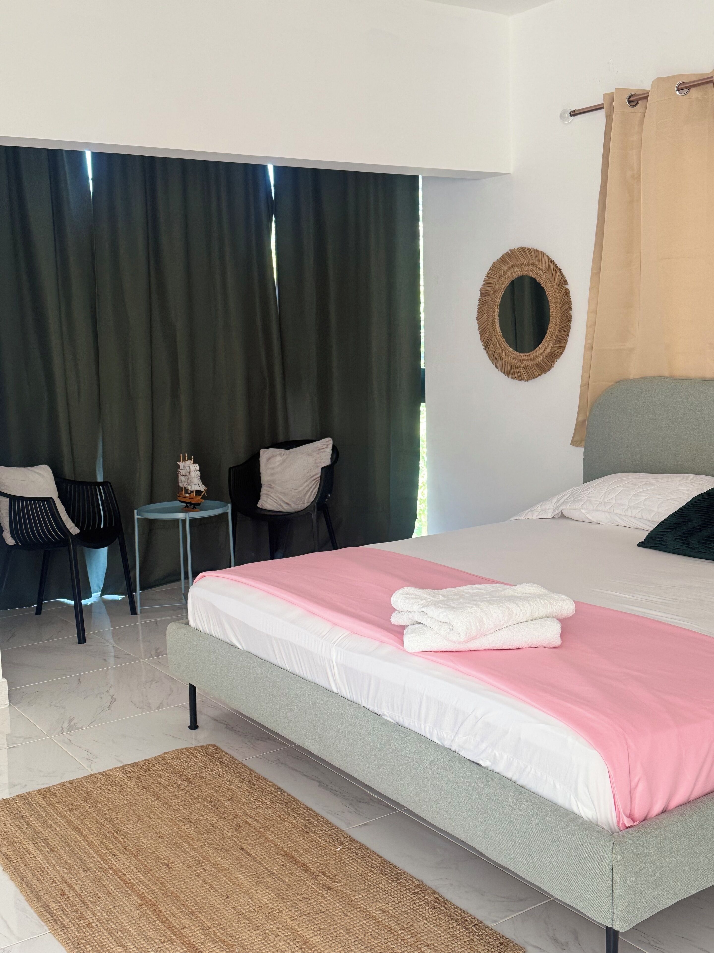 Une chambre d’hôtel moderne avec un grand lit, une petite table, deux chaises, un miroir rond et une vue sur l’extérieur à travers des rideaux sombres.
