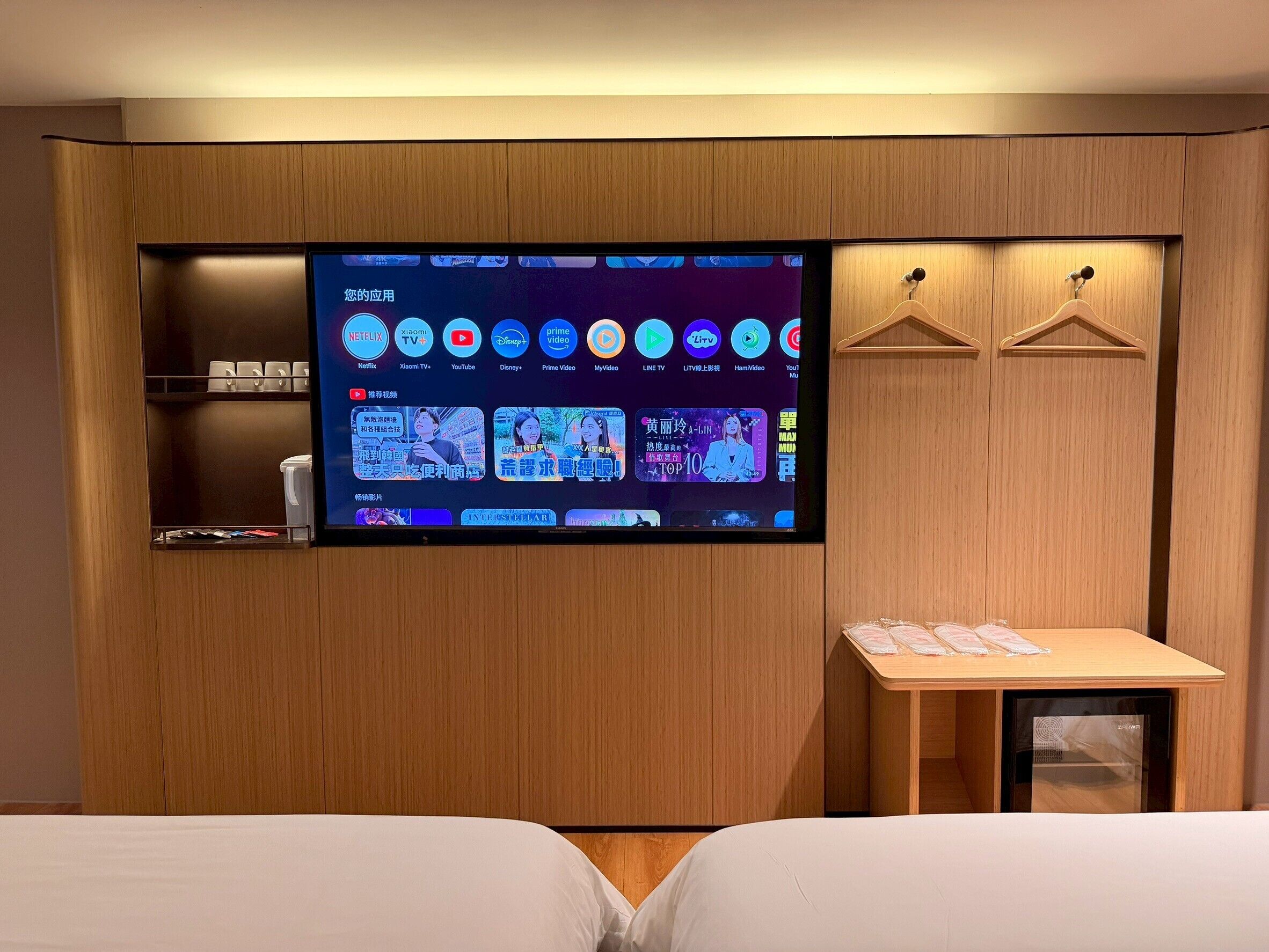 Une chambre d’hôtel dotée d’un grand téléviseur fixé au mur, qui diffuse différents services de streaming, d’une armoire en bois, d’une petite table avec un plateau et d’un lit avec du linge de lit blanc.