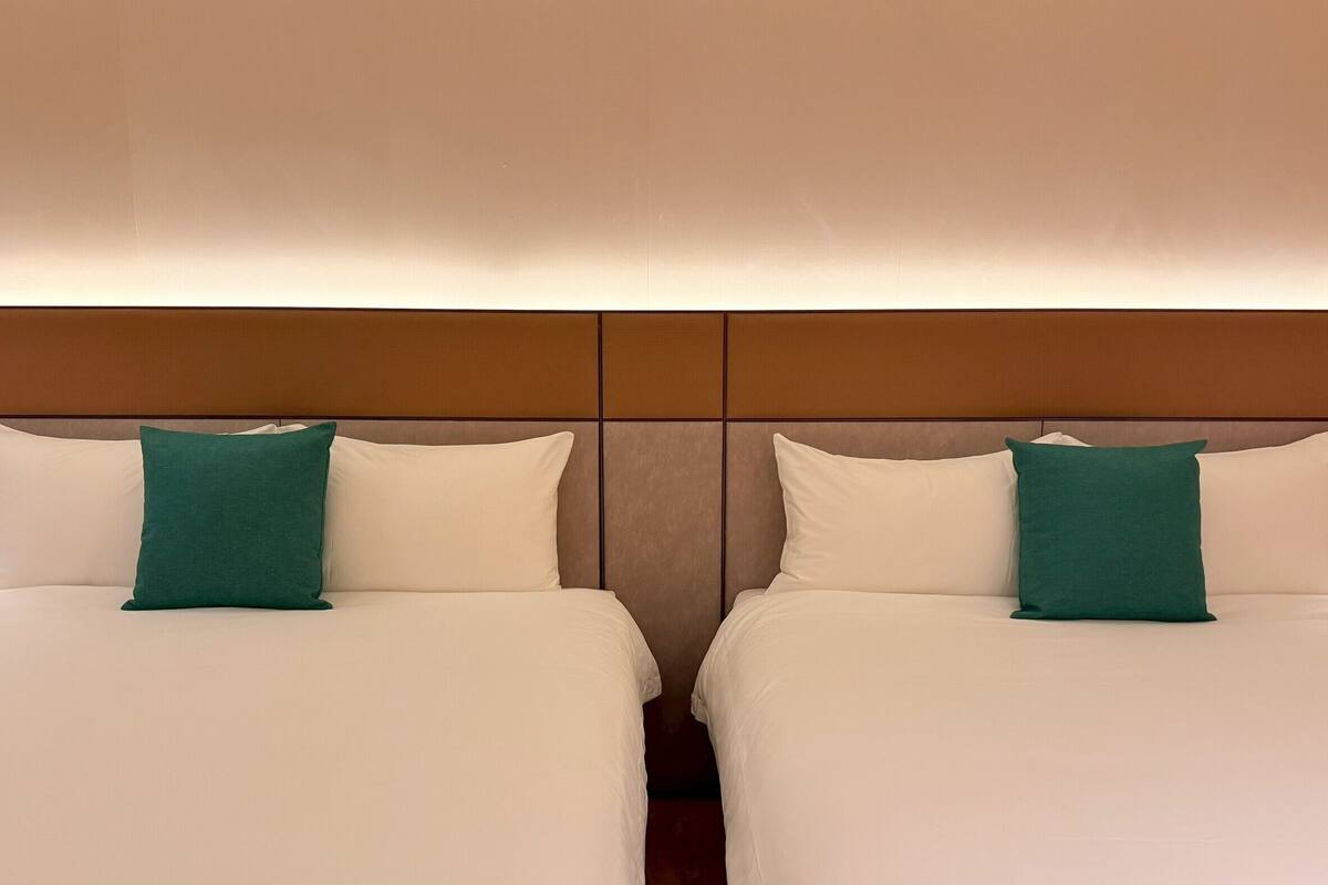 Dos camas con ropa de cama blanca y almohadas verdes, un cabecero de madera y una pared en tonos neutros.