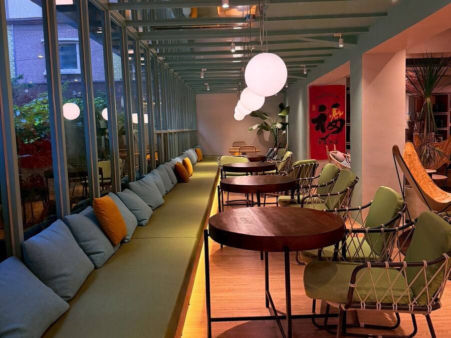 A modern interior with a long bench, round tables, wicker chairs, and a ceiling with exposed beams and hanging lights.