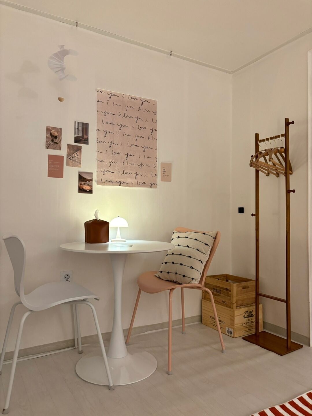 A room with a white table, a white chair, a wooden coat rack, and a wall with a large poster and several smaller photos.