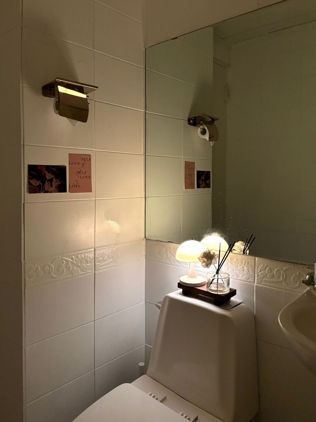 A bathroom with a white sink, a mirror, and a wall-mounted light fixture. There are decorative notes on the wall and a small table with a lamp and reed diffuser.