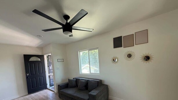 A spacious room with a ceiling fan, a dark-colored sofa, a window, and wall-mounted decorative items. - Cozy Home Minutes to Beach Cities! (Paramount)
