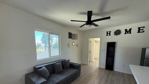 A modern living room with a dark grey sofa, a ceiling fan, a large window, and a 'HOME' sign on the wall. - Cozy Home Minutes to Beach Cities! (Paramount)