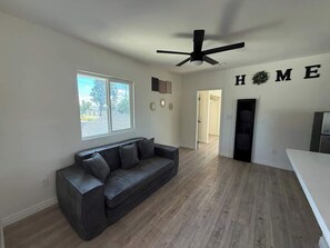 A modern living room with a dark grey sofa, a ceiling fan, a large window, and a 'HOME' sign on the wall. - Cozy Home Minutes to Beach Cities! (Paramount)
