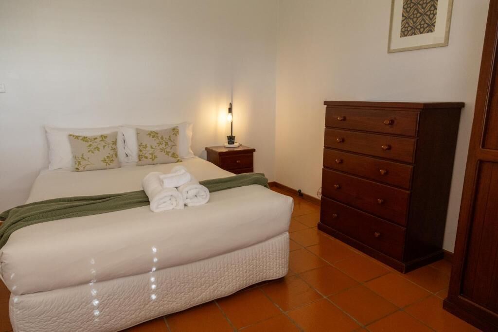 A neatly made bed with a green throw, a bedside table with a lamp, and a wooden dresser.