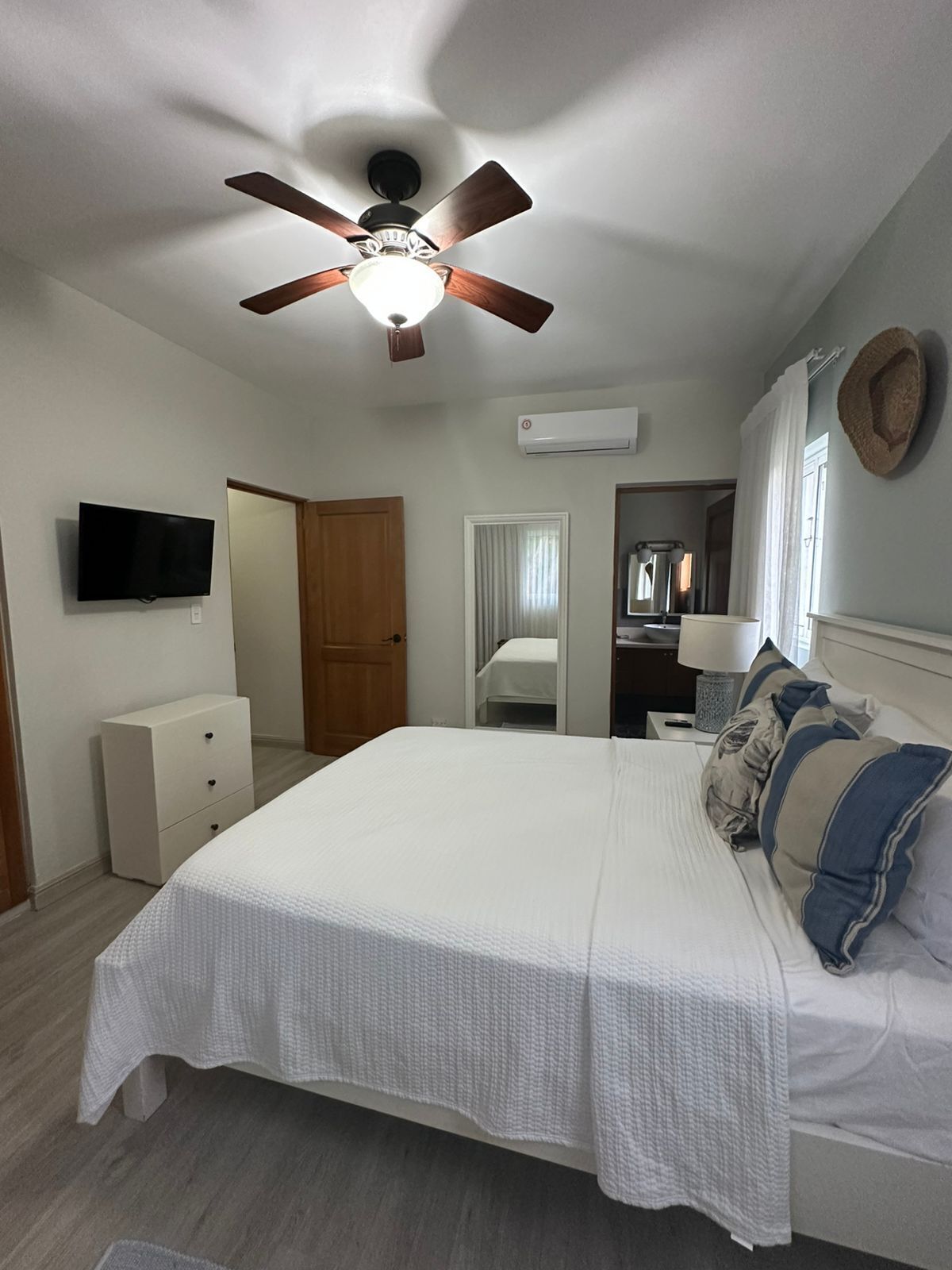 A bedroom with a bed, a ceiling fan, a TV, a wardrobe, a mirror, and an air conditioning unit.