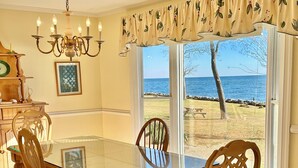 A dining room with a glass table and wooden chairs, a chandelier, a view of the ocean, and a dining table set with plates and a pitcher.