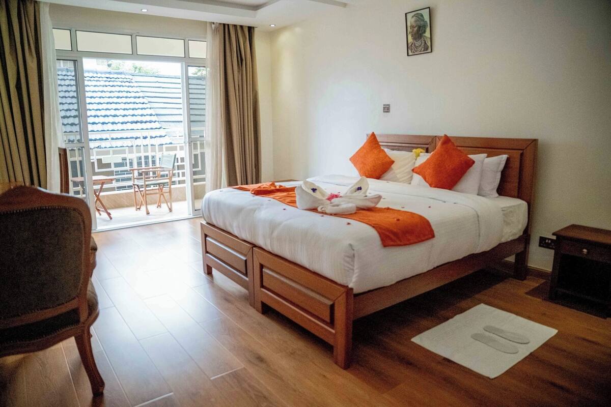 Habitación de hotel con cama de madera, ropa de cama blanca, almohadas naranjas, una toalla blanca y una mesita de noche de madera.