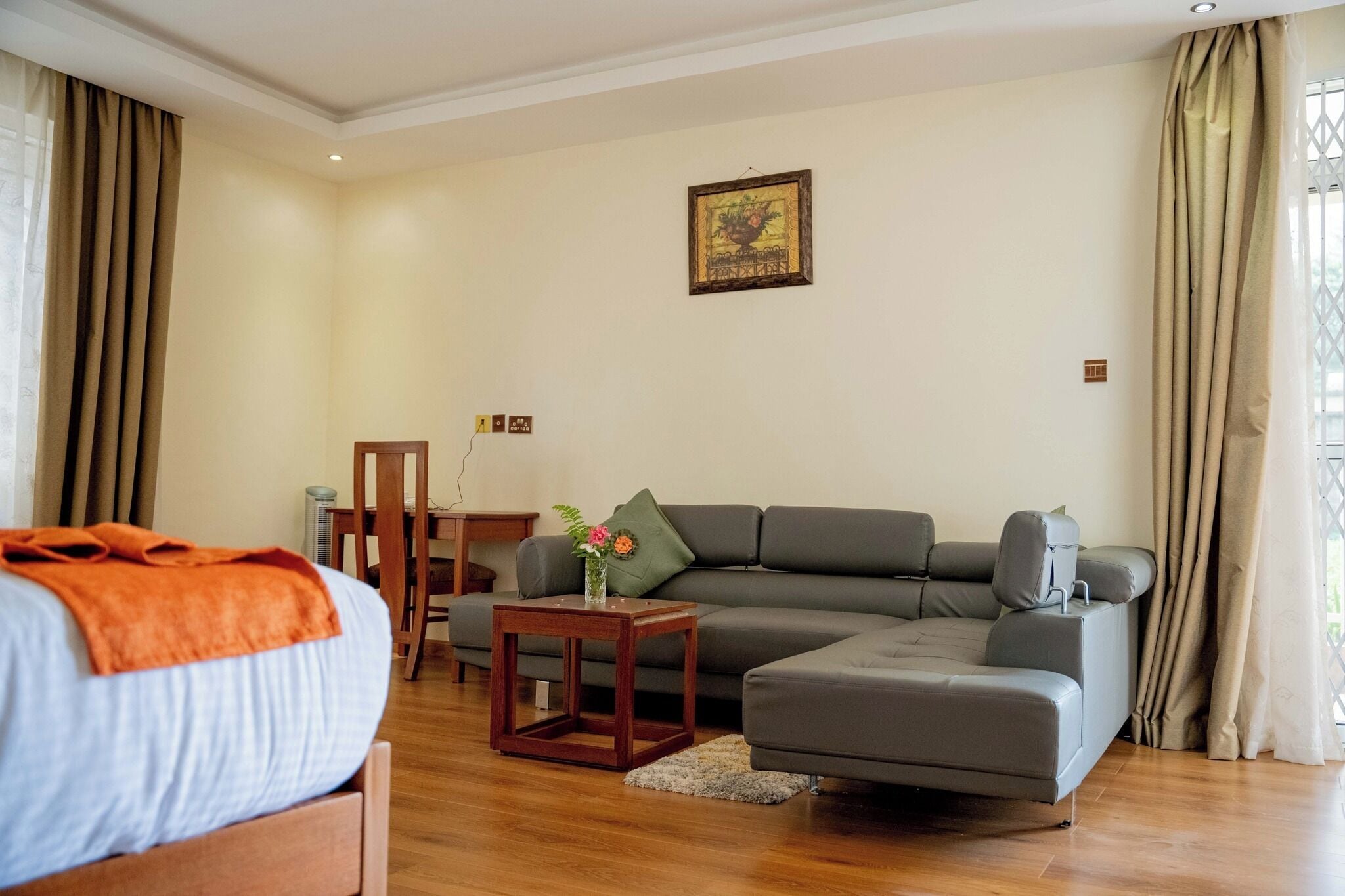 A modern living room with a grey sofa, a wooden side table with a vase of flowers, and a bed with an orange blanket.