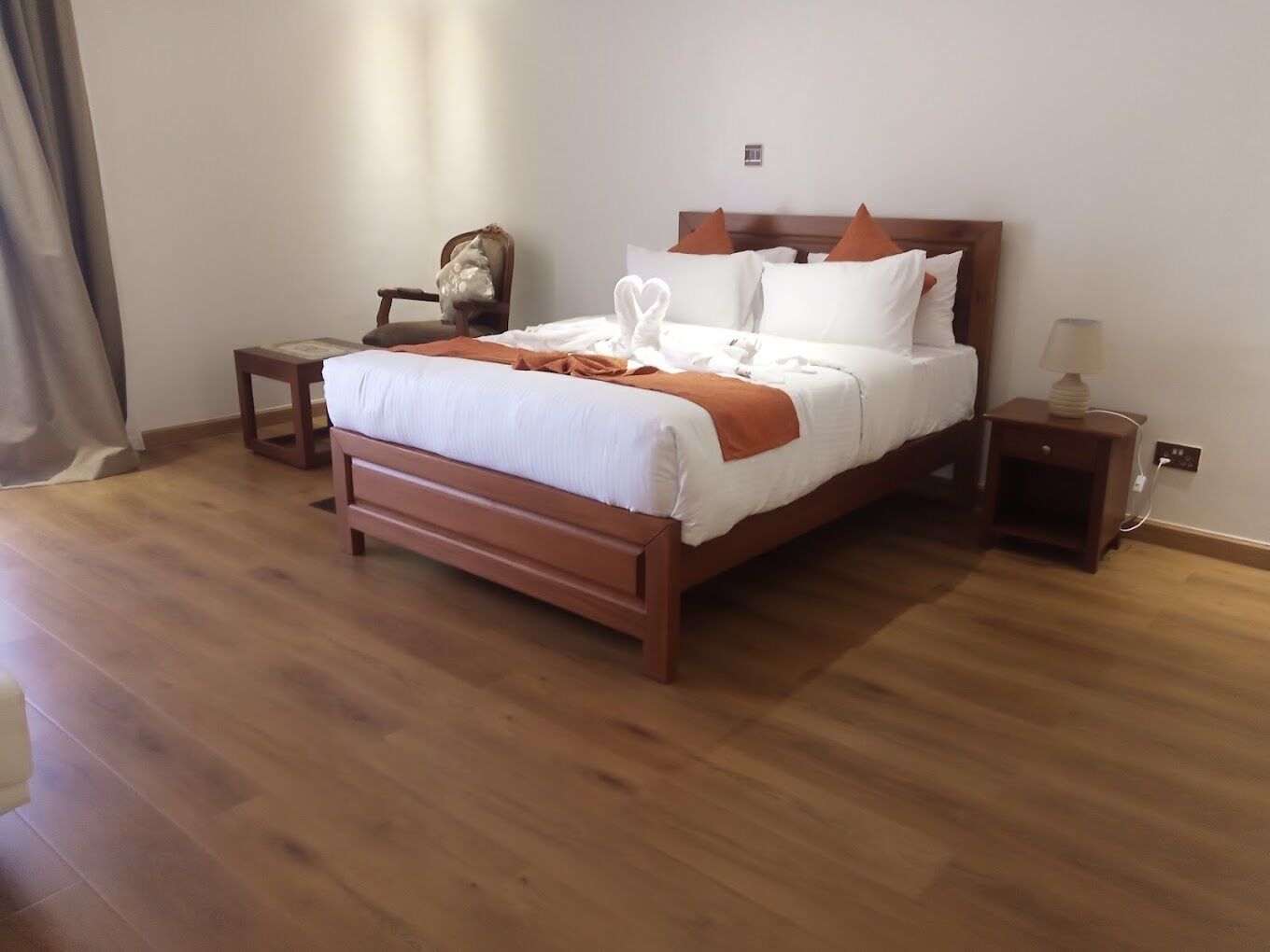 Una cama bien hecha con ropa de cama blanca y naranja, una mesita de noche de madera con una lámpara y una pequeña silla de madera.