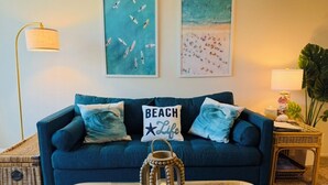 A living room with a blue sofa, a coffee table with decorative items, and wall art depicting beach scenes. - Ocean view, updated bright & relaxing beach condo close to it all! (Oceanside)