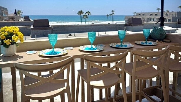 A terrace with a view of the beach, featuring a table with chairs and a table umbrella. - Ocean view, updated bright & relaxing beach condo close to it all! (Oceanside)