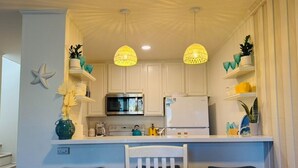 A compact kitchen with white cabinets, a microwave, and a refrigerator. There are two pendant lights, a starfish decoration, and various kitchen items on the shelves. - Ocean view, updated bright & relaxing beach condo close to it all! (Oceanside)