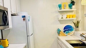 A compact kitchen with white cabinets, a microwave, a stove, and a sink. There are decorative items on the shelves and a small rug on the floor. - Ocean view, updated bright & relaxing beach condo close to it all! (Oceanside)
