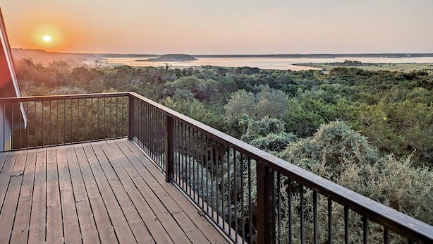 Une terrasse en bois avec une rambarde en métal, d’où l’on peut admirer un paysage verdoyant et une étendue d’eau au loin.