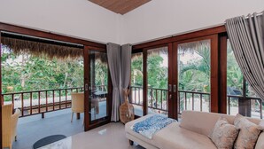 A spacious room with a large sliding glass door leading to an outdoor area with a thatched roof and a view of greenery. There is a sofa with patterned cushions and a wicker chair. - 28 Degrees Lembongan (Lembongan Island)