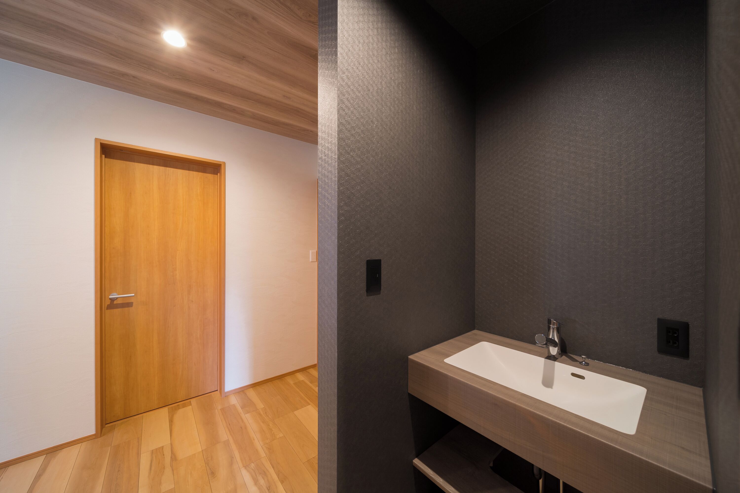 Un baño moderno con una puerta de madera, un lavabo blanco y una pared oscura con textura.