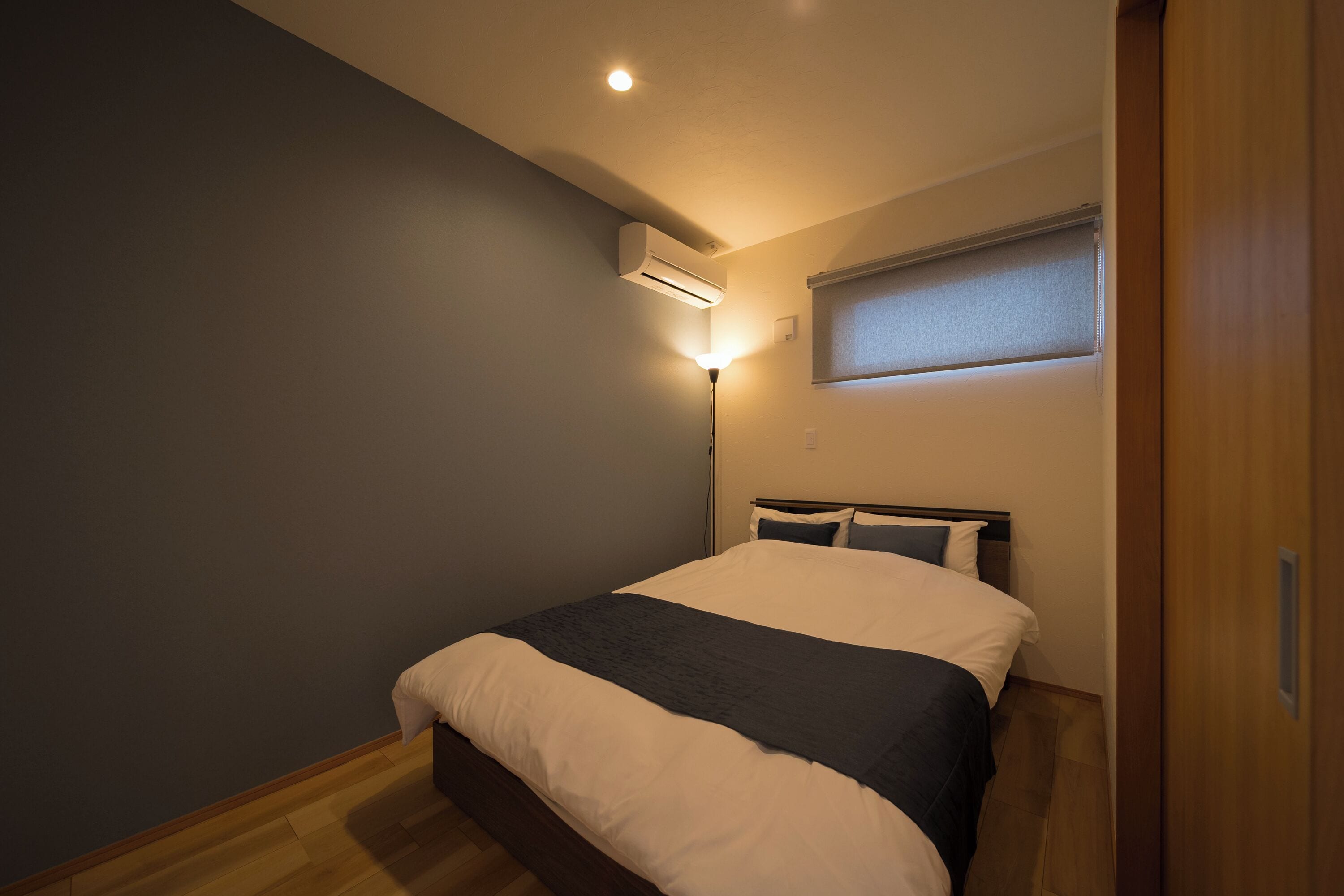 Un dormitorio con cama, mesita de noche, aire acondicionado, lámpara de pared y ventana con persianas.