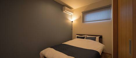 Un dormitorio con cama, mesita de noche, aire acondicionado, lámpara de pared y ventana con persianas.
