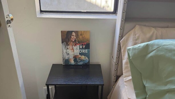Een slaapkamer met een bed, nachtkastje met een boek, een raam en een deur.