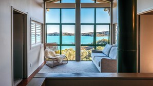 A spacious room with a large window offering a view of a lake and hills. There is a wicker chair and a sofa with a view. - Spinnaker Watch - Waiheke Island (Waiheke Island)