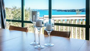 A table with two clear glass wine glasses and two white candles, set against a backdrop of a scenic view through large windows. - Spinnaker Watch - Waiheke Island (Waiheke Island)