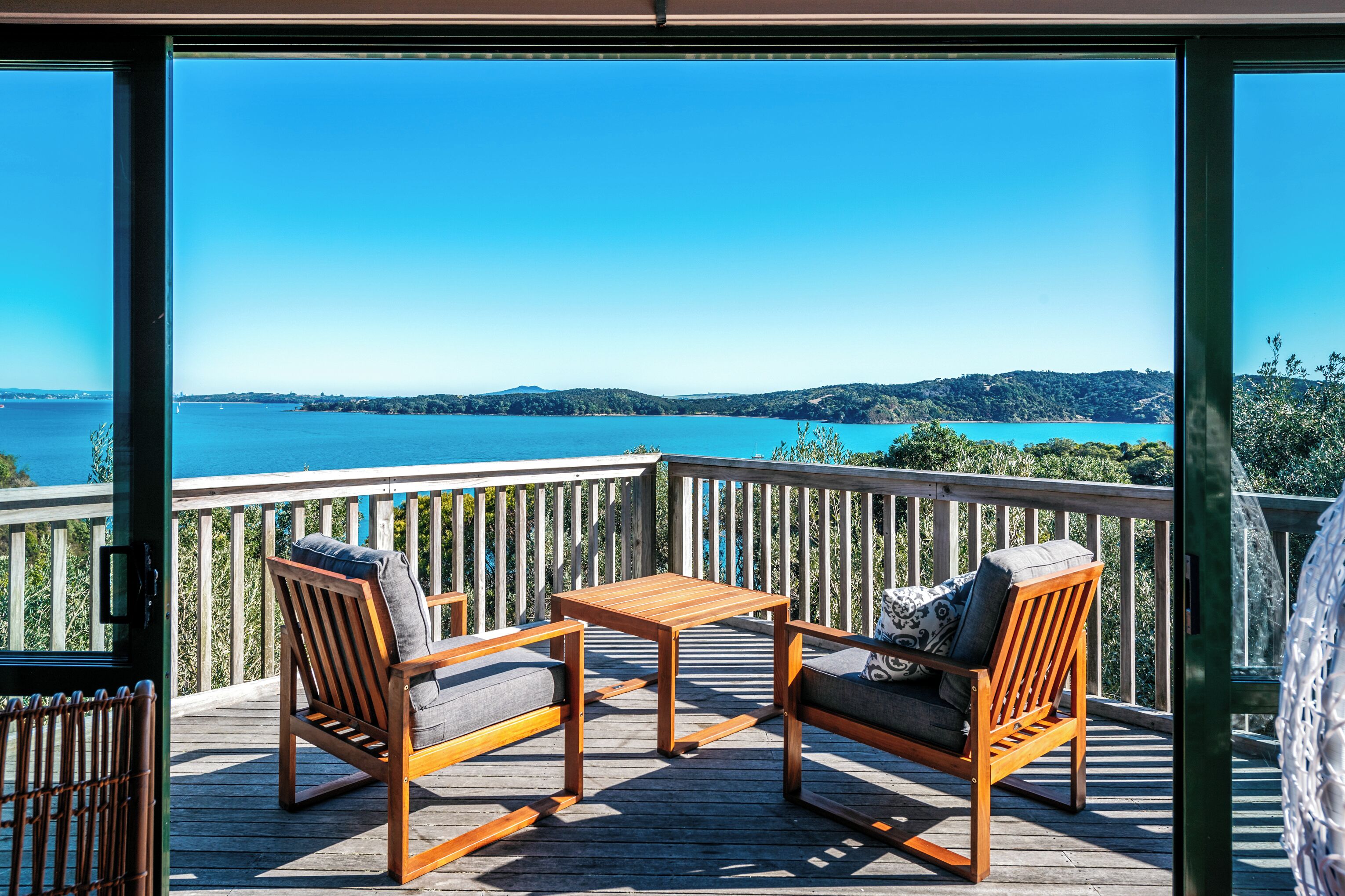 Eine Holzterrasse mit zwei Stühlen und einem Tisch, mit Blick auf klares blaues Wasser und ferne Hügel.