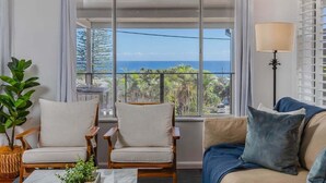 A room with a large window offering a view of the ocean, two wooden chairs, a sofa, a lamp, and a small plant.