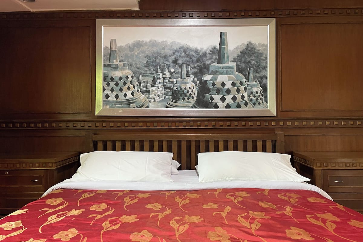 A hotel room with a large bed, two white pillows, a red floral bedspread, and a framed painting of traditional architecture on the wall.