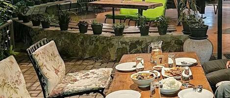 Un restaurante con una mesa de madera dispuesta para una comida, rodeada de sillas. Hay plantas en macetas y luces colgantes.