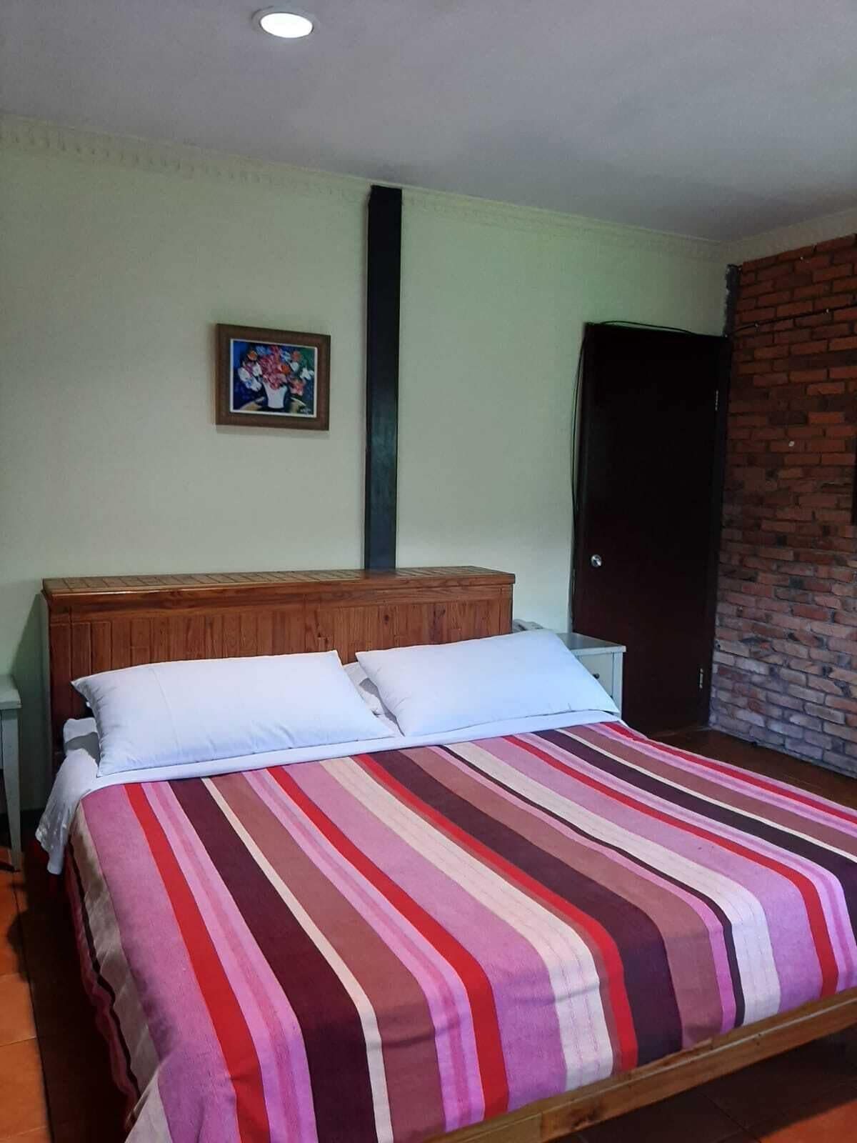 Habitación con una cama individual, cabecera de madera, colcha a rayas, mesita de noche pequeña, un aparador y una fotografía enmarcada en la pared, todo sobre un muro de ladrillo.
