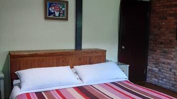 Habitación con una cama individual, cabecera de madera, colcha a rayas, mesita de noche pequeña, un aparador y una fotografía enmarcada en la pared, todo sobre un muro de ladrillo.